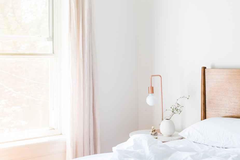 Copper light fixture on a bedside table next to a wooden headboard and soft white bedding in a bright bedroom.