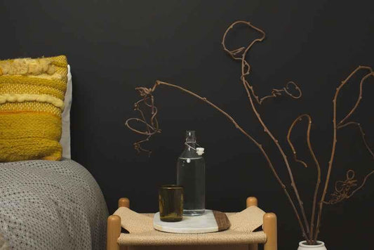 A cozy bedroom corner featuring a textured blanket, a decorative pillow, a glass bottle, and dried branches on a small table against a dark wall.