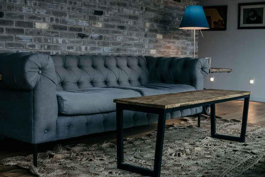 A cozy living room scene featuring a tufted gray sofa, a wooden coffee table, and a blue lamp against a brick wall.